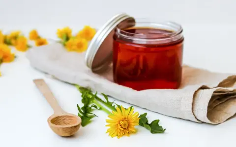 Słoik z bursztynowym syropem z mniszka 500 kwiatów, obok drewniana łyżeczka i świeże kwiaty mniszka.