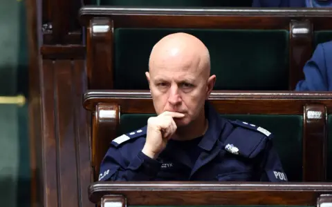 Łysy mężczyzna w mundurze policji siedzi w drewnianej ławie, zamyślony.