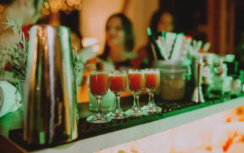 Cztery kieliszki z czerwonym drinkiem stoją na barze, gotowe do podania na weselu. W tle widać gości i dekoracje.