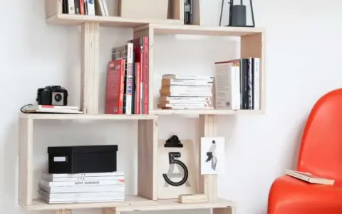Bibliothèque DIY en bois clair, remplie de livres, d'une caméra vintage et d'une boîte noire. Une chaise rouge vif est visible à droite.