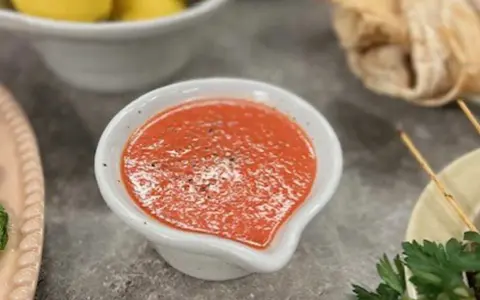 En skål med en röd, kryddig sås, kanske en pico de gallo, står på ett grått bord. Bredvid syns gula citroner och bröd.