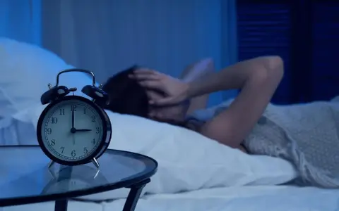 Eine Frau hält sich im Bett die Ohren zu, während ein Wecker auf 3 Uhr steht. Warum wache ich immer um 3 Uhr auf?
