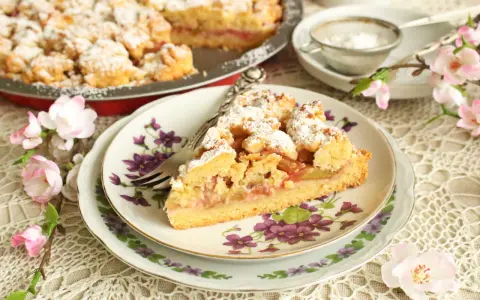 Pyszna tarta z rabarbarem, posypana cukrem pudrem, na talerzyku z kwiatowym wzorem, obok gałązka kwitnącej wiśni.