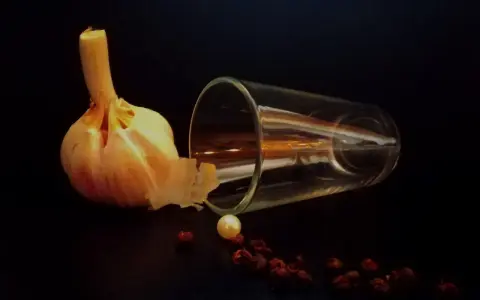 Photographie nature morte : une tête d'ail, un verre renversé, une perle et des baies de poivre rouge sur fond sombre.