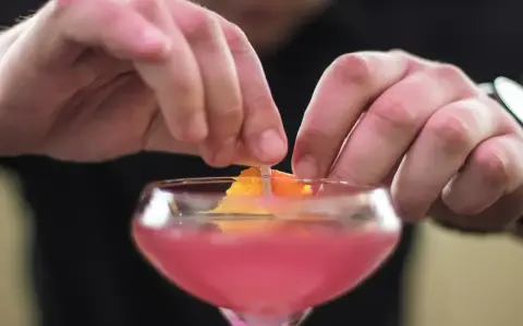 Dłonie barmana ozdabiają drinka skórką pomarańczy. To klasyczny cosmopolitan, gotowy na wieczór w wielkim mieście.