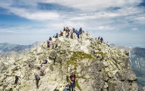 Tłum turystów na skalistym szczycie Rysów, z widokiem na Giewont w oddali.