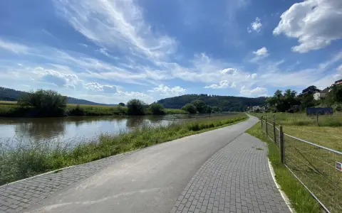 Ein asphaltierter Weg säumt den Fluss, ideal für eine Weser-Radweg Rundtour. Grüne Ufer und ein blauer Himmel mit Wolken laden zum Radfahren ein.