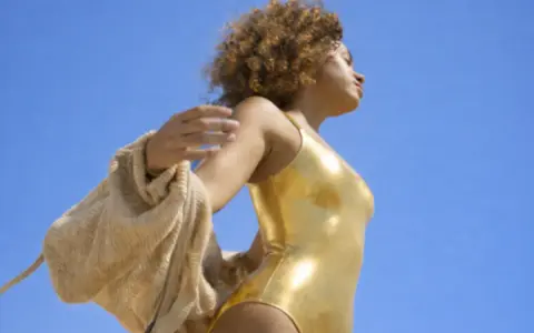 Femme en maillot doré, bras écartés, profitant du soleil. Une belle façon de préparer sa peau au soleil.