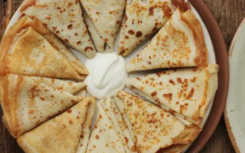 Piatto di crespelle ripiene, piegate a spicchi, con una generosa porzione di panna acida al centro.
