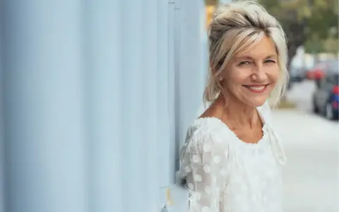 Donna sorridente appoggiata a un muro blu. Potrebbe beneficiare di acido folico in menopausa per il suo benessere.