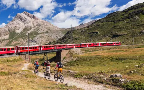 Treno rosso attraversa un ponte su un paesaggio alpino mozzafiato. Tre ciclisti pedalano su un sentiero sottostante, godendosi la vista della ferrovia del Bernina.