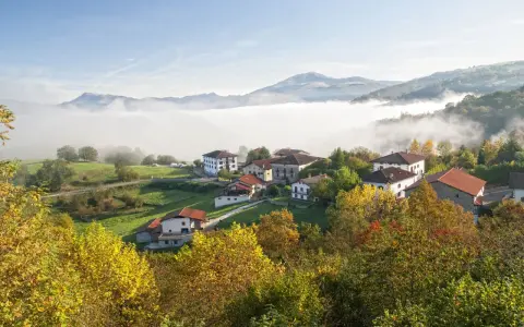 Pueblo en el **valle del Baztan** cubierto por niebla matutina, con casas blancas y tejados rojos entre árboles otoñales.