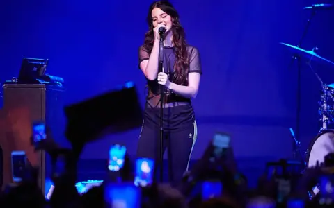 Lana Del Rey, w czarnej, prześwitującej bluzce i sportowych spodniach, śpiewa na scenie. Fani z telefonami tworzą tłum. To jej lana del rey opener.
