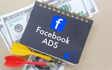 Notatnik z logo Facebook i napisem "Facebook ADS" leży na banknotach dolarowych. Obok znajdują się rzutki, symbolizujące celne reklamy mobilne.