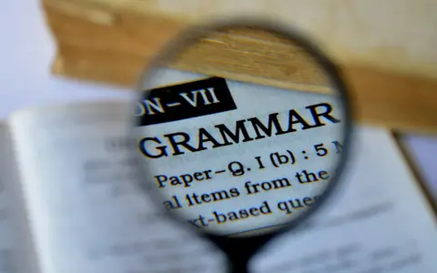 Une loupe examine le mot "GRAMMAR" dans un livre. Pour maîtriser la langue, il faut bien receler la conjugaison.