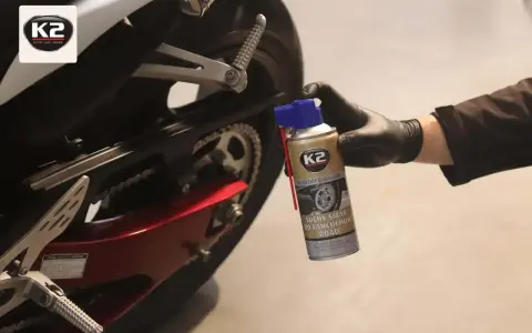 Ręka w czarnej rękawiczce aplikuje suchy smar do łańcucha motocyklowego K2. Idealny wybór, by zadbać o napęd.