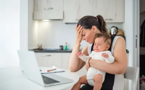Zmęczona mama z płaczącym niemowlakiem na rękach, zastanawia się, przebodźcowanie niemowlaka ile trwa. Obok laptop i dokumenty.