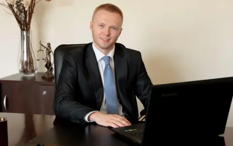 Mężczyzna w garniturze siedzi przy biurku z laptopem, obok statuetka Temidy.