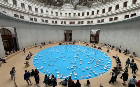 A circular room with a frescoed ceiling, featuring a bright blue pool dotted with white ceramic bowls. Visitors observe this installation, part of the François-Henri Pinault art collection.