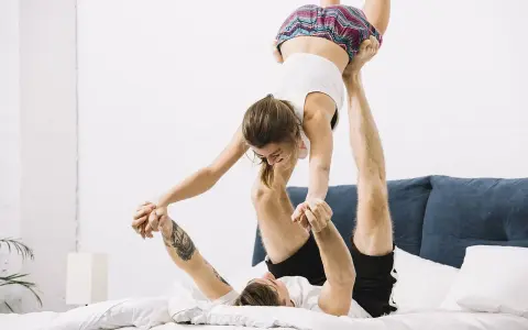 Para ćwiczy akrobatykę na łóżku. Kobieta w powietrzu, mężczyzna podtrzymuje ją nogami, tworząc pozycję seksualną pająk.