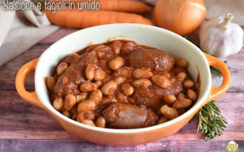 Piatto fumante di salsiccia e fagioli in umido, con carote, cipolla e aglio sullo sfondo.