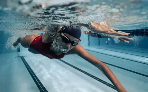Une femme en maillot de bain rouge nage en crawl, lunettes de natation sur le nez. Sa **natation transformation physique femme** est visible sous l'eau.