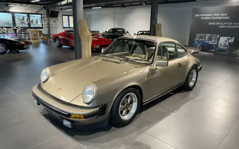 Ein goldener Porsche 911 SC 3.0 steht in einer Ausstellungshalle, flankiert von anderen Sportwagen.