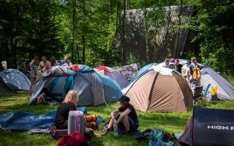 Ludzie rozbijają namioty na polu namiotowym, gotowi na festiwal. W tle widać budynek, a wokół drzewa. To idealne miejsce na nocleg podczas Pyrkonu, gdzie każdy znajdzie swoje miejsce i pyrkon tokeny.