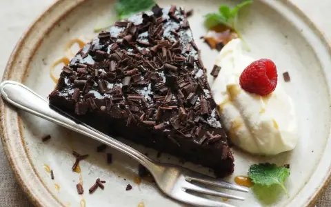 En bit glutenfri kladdkaka toppad med chokladspån, serverad med vispad grädde, hallon och mynta.