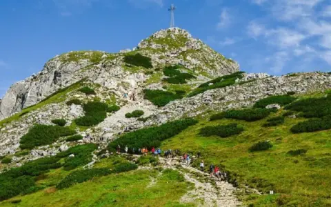 Grupa turystów wchodzi kamienistym szlakiem na zielone zbocze góry z krzyżem na szczycie.