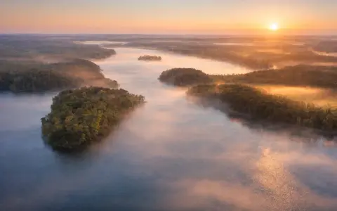 Mglisty poranek nad jeziorem Mukrza. Słońce wschodzi nad lasami, tworząc złote refleksy na wodzie i unosząc się nad mgłą.