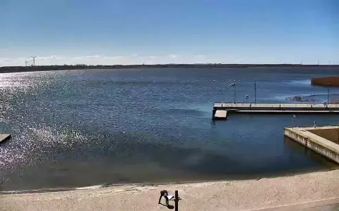 Widok z jeziorsko kamera na żywo. Na piaszczystej plaży dwie osoby, w tle spokojne jezioro, pomost i wiatraki.