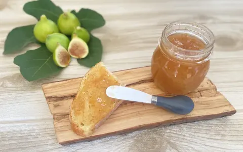 Fetta di pane con marmellata di fichi, accanto a un vasetto e a fichi freschi su un tagliere di legno.