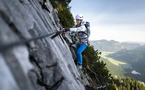 Kobieta w kasku i uprzęży pokonuje via ferrata, chwytając się stalowej liny. W tle rozciąga się malownicza górska dolina.