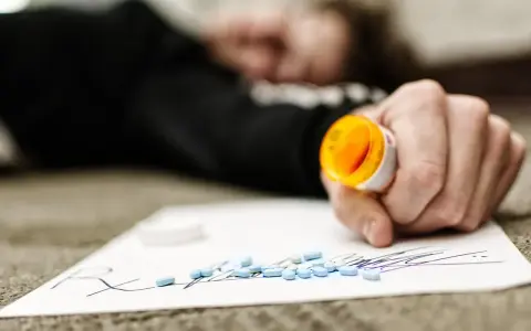 Dłoń trzymająca otwartą butelkę z lekami, obok rozsypane tabletki na kartce z napisem "Rx".