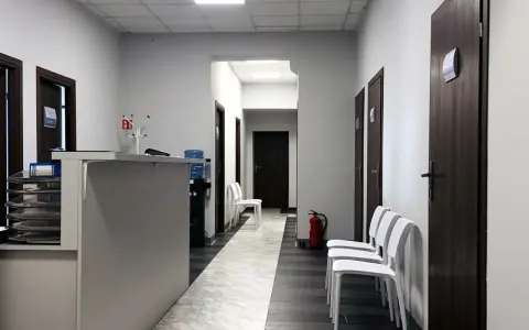 Korytarz kliniki medycznej z recepcją, drzwiami i białymi krzesłami.