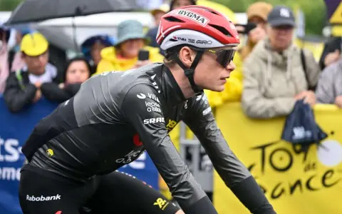 Trine Vingegaard, a cyclist in a grey and black racing suit, rides in the rain. Spectators watch from behind a yellow barrier with "Tour de France" branding.