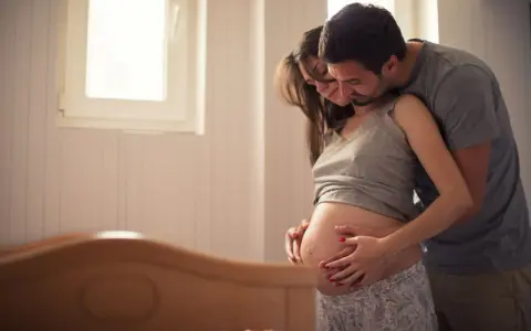 Ein Mann umarmt seine schwangere Frau, die ihren Bauch streichelt. Sie warten gespannt auf ihr Baby. Senkwehen ab wann?