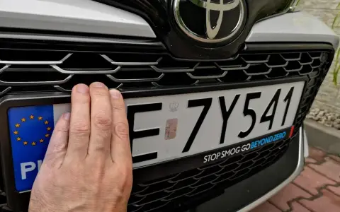 Ręka zasłania część tablicy rejestracyjnej. Upewnij się, że masz aktualne dokumenty, by uniknąć kary za nieprzerejestrowanie samochodu.
