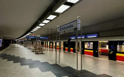 Pusta platforma stacji metra M1 Politechnika z pociągiem i tablicami kierunkowymi do Kabat i Młocin.