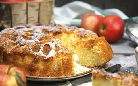 Pyszne ciasto z jabłkami, posypane cukrem pudrem, z kilkoma jabłkami w tle. Idealne na jesienne popołudnie.