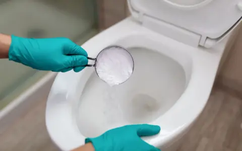 Hands in teal gloves pour baking soda into a toilet bowl, a method for how to unclog toilet without plunger.