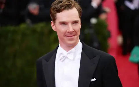 Benedict Cumberbatch w czarnym smokingu i eleganckim białym krawacie, z uśmiechem pozuje na czerwonym dywanie.