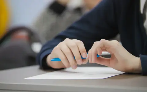 Uczeń pisze wypracowanie, przygotowując się do matury 2026. Na biurku biała kartka i niebieski długopis. Lektury obowiązkowe czekają.