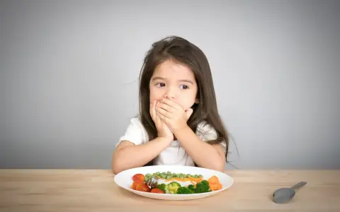Kleines Mädchen bedeckt Mund, schaut auf Teller mit Gemüse. Manche kinder essen schnell, aber dieses hier scheint zu überlegen.