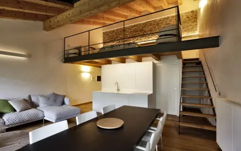 Modern living space with a loft bedroom accessible by stairs. The **soppalco soffitto basso** features a metal railing and a stone wall.