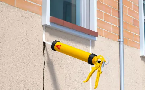 Pistola de calafateo amarilla aplicando sellador blanco para como tapar grietas en la pared exterior de estuco junto a una ventana.