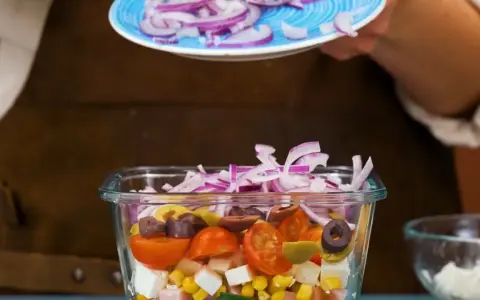 En person lägger rödlök på en pastasallad med kassler, majs, tomater, oliver och fetaost.