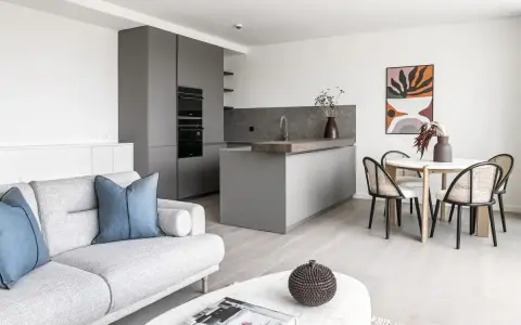 Salón moderno y cocina integrada, ideal para amueblar piso de alquiler. Sofá gris, mesa redonda con sillas de ratán y arte abstracto.