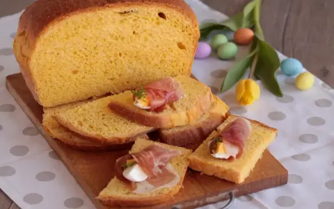 Fetta di torta al formaggio umbra ricetta originale, guarnita con uovo sodo e prosciutto crudo, su tagliere di legno, con uova colorate e tulipano.